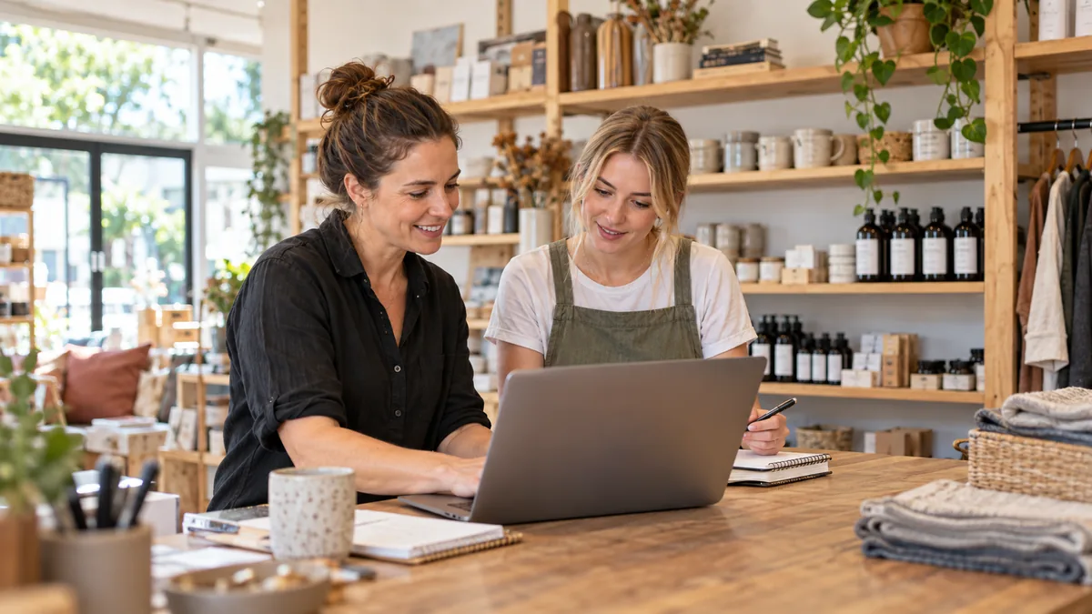 Small retail owner working with staff member on website content at shop desk, notebooks and product shelves.